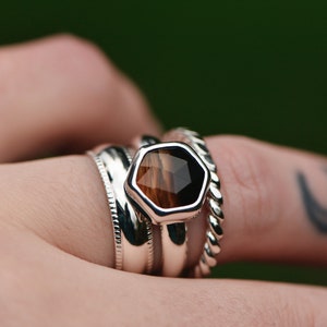 Può includere: Un anello d'argento con una pietra preziosa esagonale marrone e nera incastonata al centro. L'anello è impilato con altre due bande d'argento.
