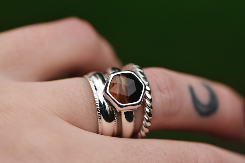 May include: A silver ring with a hexagonal brown and black gemstone set in the center. The ring is stacked with two other silver bands.