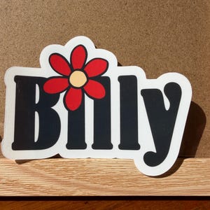 May include: A die-cut sticker featuring the name Billy in bold black lettering with a white outline. A red flower with a pale yellow center is positioned above the letter i, replacing the dot. The sticker has a white border throughout.