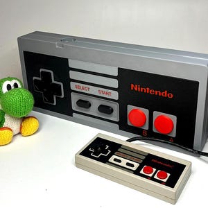 Pode incluir: Uma caixa cinza em forma de controle Nintendo com um controle menor e uma figura de Yoshi de crochê verde e branco. O controle grande tem botões vermelhos e o logotipo da Nintendo. O controle menor é bege.