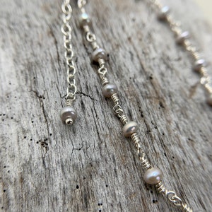May include: A silver chain necklace with small, white pearls strung on it. The chain has a delicate, wire-wrapped design and a small pearl dangle at the end.