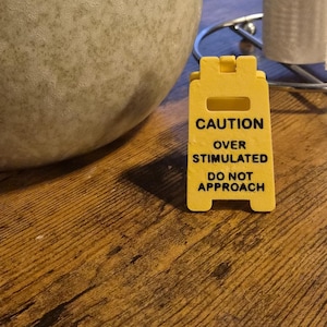 Könnte beinhalten: Ein gelbes Warnschild mit dem Text "CAUTION OVER STIMULATED DO NOT APPROACH" steht auf einer Holzoberfläche. Eine große, gesprenkelte, beige Keramiktasse und ein Papierhandtuchhalter aus Chrom mit einer Rolle weißer Papierhandtücher sind ebenfalls zu sehen.