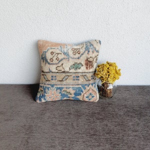 May include: A decorative throw pillow with a vintage floral pattern in shades of blue, pink, and beige. The pillow is sitting on a brown couch with dried flowers in a glass vase.