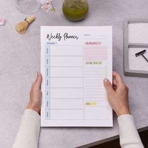 Op de afbeelding: Een witte weekplanner met een lichtblauw, roze en geel kleurenschema. De planner heeft secties voor "Schedule", "Top Priorities", "Weekly Task List" en "Notes".