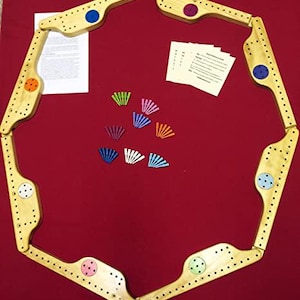 May include: A wooden cribbage board game set with a hexagonal playing surface and pegs in various colors. The board is made of light wood with a natural finish. The pegs are arranged in a fan shape on the table.