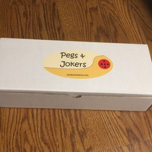 May include: A white rectangular box with a tan and red oval sticker that reads "Pegs & Jokers" with the website address candiwoodtechs.com. The box is sitting on a wooden surface.