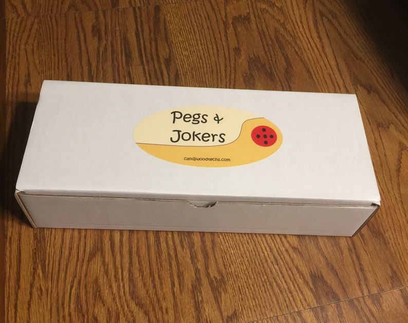 May include: A white rectangular box with a tan and red oval sticker that reads "Pegs & Jokers" with the website address candiwoodtechs.com. The box is sitting on a wooden surface.