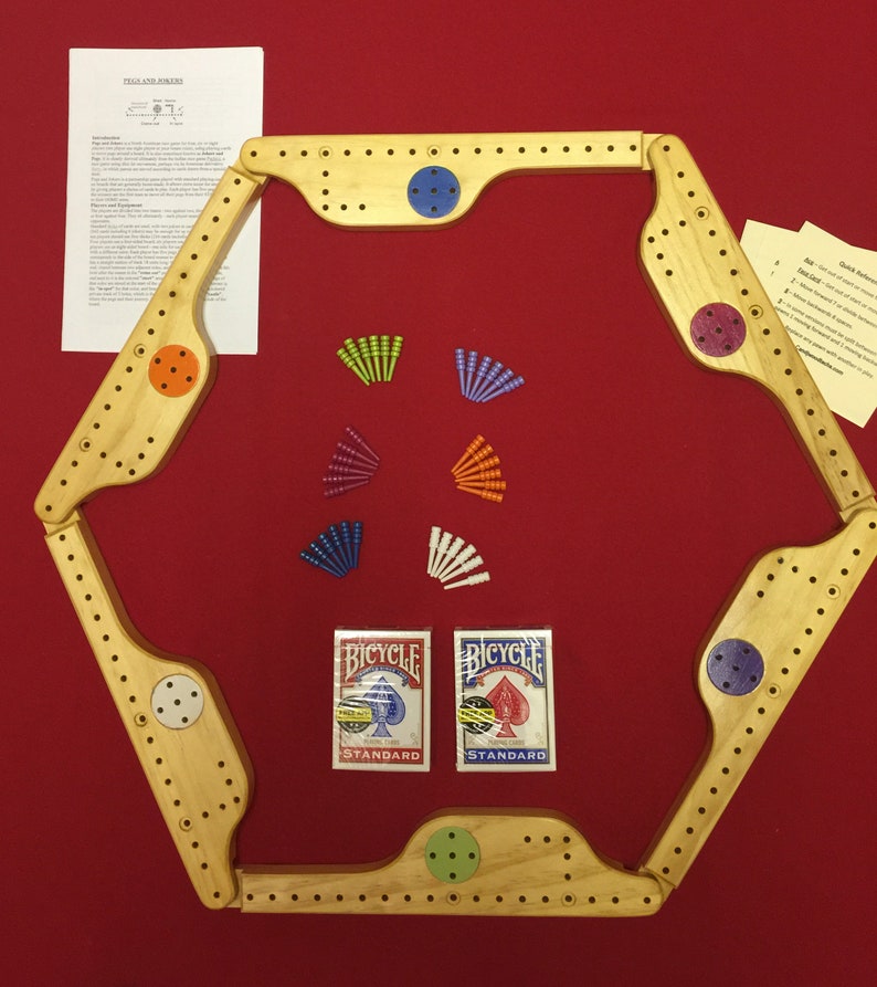 May include: A wooden Pegs and Jokers board game on a red surface. The game includes a wooden hexagonal board, coloured pegs, and two decks of Bicycle playing cards. Instructions are included.