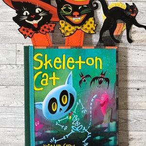 May include: A hardcover book titled Skeleton Cat by Kristyn Crow with illustrations by Dan Krall, featuring a blue cat skeleton on the cover. Three decorative cat-themed bookmarks are positioned above the book, including two with hats and one black cat silhouette.