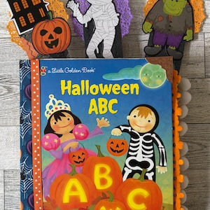 May include: A Little Golden Book titled Halloween ABC featuring a child in a princess costume and a child in a skeleton costume with pumpkins. The book is decorated with orange ribbon, white pom-poms, and three Halloween-themed paper cutouts.