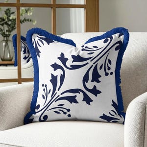 May include: A square throw pillow featuring a white fabric base with a dark blue floral pattern. The pillow is finished with a dark blue fringe trim along all four edges, creating a textured border around the central botanical design.