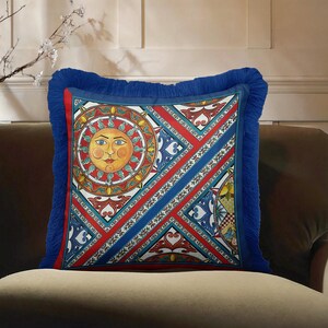 May include: A square decorative cushion featuring a vibrant, multi-coloured pattern with a central sun face motif. The design includes geometric sections in red, white, and blue, finished with a thick, deep blue fringe border on all four sides.