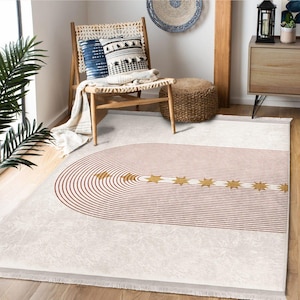 May include: A cream-colored area rug with a geometric design. The rug features a series of concentric, reddish-brown curved lines and a straight line with gold star accents. The rug is fringed on the edges and is placed on a wooden floor.
