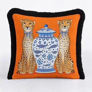 May include: A square throw cushion with a vibrant orange background featuring two cheetahs wearing blue and white striped neck ribbons, flanking a central blue and white porcelain ginger jar with fish motifs, finished with a black fringe border.