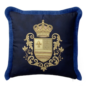 May include: A square navy blue throw pillow featuring a gold embroidered crest with a crown and a fleur-de-lis. The pillow is finished with a matching navy blue fringe trim along all four edges, creating a decorative border on the dark fabric.