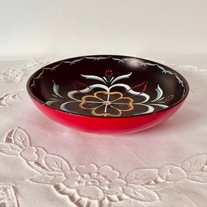 May include: A small, red and black wooden bowl with a hand-painted floral design. The bowl has a black interior and a red exterior with a white, yellow, and blue floral design.