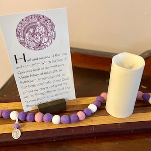 May include: A wooden prayer tray with a purple, pink, and white felt rosary, a small silver charm, and a white candle. The tray has a dark wood border and a light wood center. A card with a prayer to the Blessed Mother is propped up on the tray.