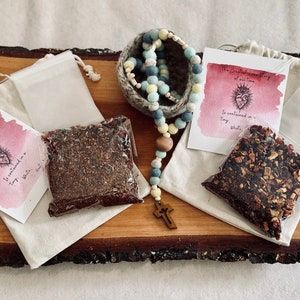 May include: A wooden serving board displays tea-related items. Two clear bags of loose tea, two small muslin bags, and two cards with text and a heart illustration are arranged around a rosary with a wooden cross. A small, woven basket holds the rosary.