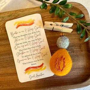 May include: A card with a prayer to the Holy Spirit, written in a cursive font, with a yellow, orange, and red watercolor background. The card is next to a small, orange felt ornament with a red flame design.