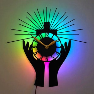 May include: A rainbow-coloured clock with black hands and a black silhouette of two hands holding the clock face. The clock face has a black background with white numbers and a gold-coloured hour and minute hand.
