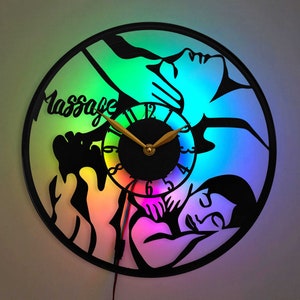 May include: A black clock with a rainbow background and a silhouette of a person receiving a massage. The clock face has a gold hour and minute hand and the words "Massage" are printed on the clock face.