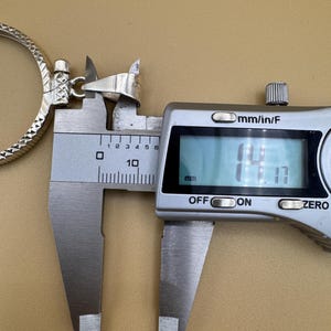 Puede incluir: Primer plano de un colgante de plata medido con un calibre digital. El calibre muestra 14,1 mm.