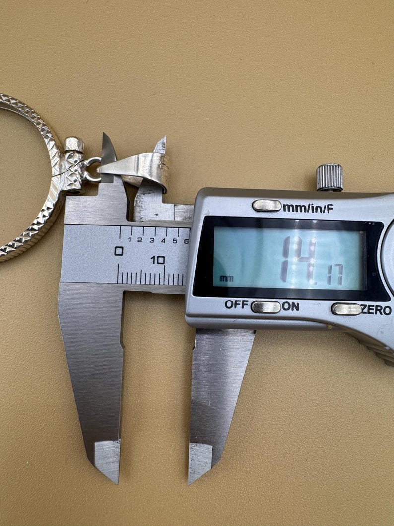 Puede incluir: Primer plano de un colgante de plata medido con un calibre digital. El calibre muestra 14,1 mm.