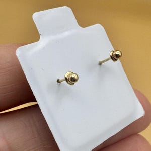 Puede incluir: Un par de peque&ntilde;os pendientes de bot&oacute;n en forma de bola dorada con cierres de rosca a juego, montados en una tarjeta de presentaci&oacute;n rectangular blanca. Los pendientes presentan un dise&ntilde;o esf&eacute;rico pulido y postes roscados.