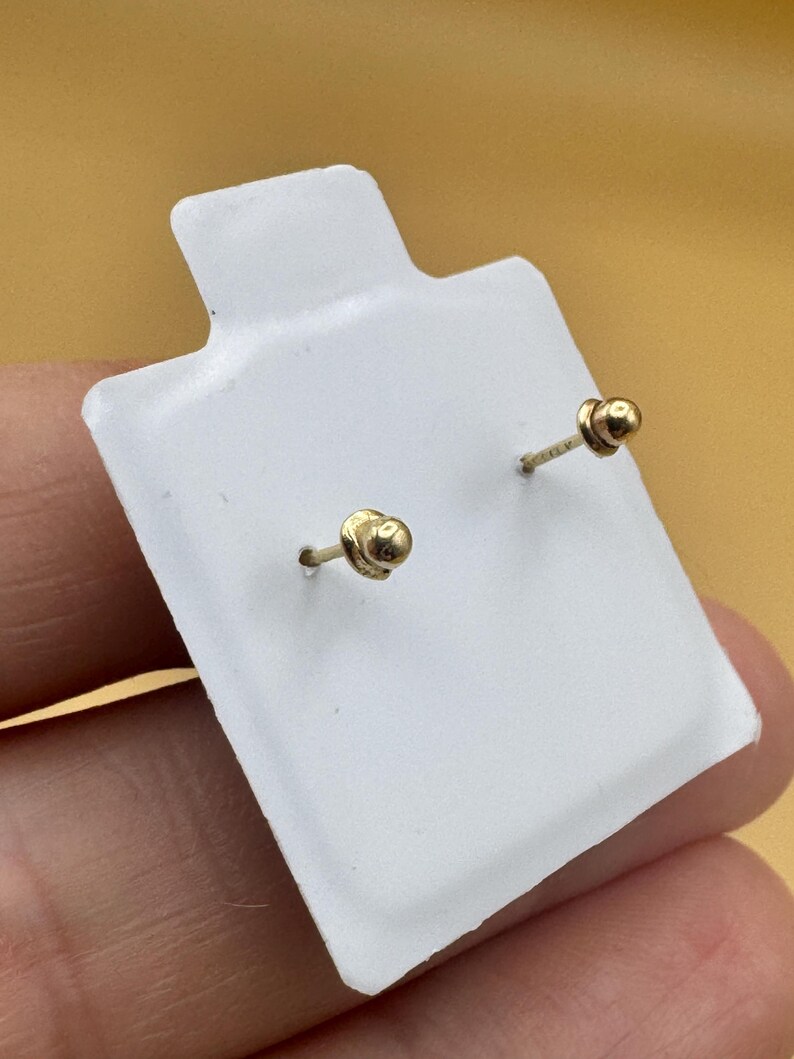 Puede incluir: Un par de peque&ntilde;os pendientes de bot&oacute;n en forma de bola dorada con cierres de rosca a juego, montados en una tarjeta de presentaci&oacute;n rectangular blanca. Los pendientes presentan un dise&ntilde;o esf&eacute;rico pulido y postes roscados.
