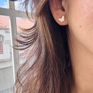 Puede incluir: Un peque&ntilde;o pendiente de bot&oacute;n con forma de mariposa, con una superficie blanca iridiscente y lisa y un borde fino de metal dorado, sujeto al l&oacute;bulo de la oreja. El dise&ntilde;o de la mariposa presenta contornos de alas delicados.