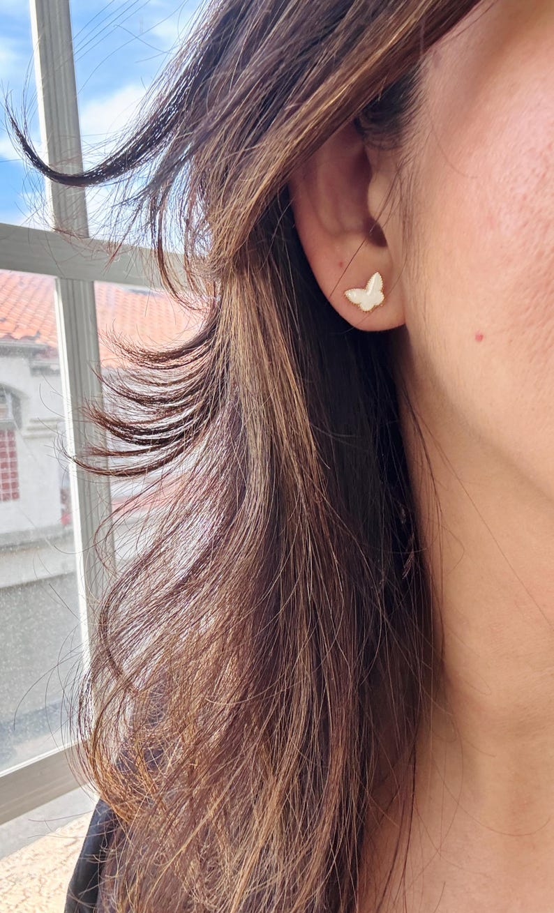 Puede incluir: Un peque&ntilde;o pendiente de bot&oacute;n con forma de mariposa, con una superficie blanca iridiscente y lisa y un borde fino de metal dorado, sujeto al l&oacute;bulo de la oreja. El dise&ntilde;o de la mariposa presenta contornos de alas delicados.