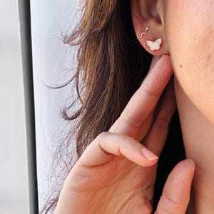 Puede incluir: Un pendiente tipo ear cuff dorado con forma de serpiente fina y sinuosa, colocado sobre un peque&ntilde;o pendiente de bot&oacute;n con forma de mariposa blanca. Las piezas de joyer&iacute;a se llevan en el l&oacute;bulo y el cart&iacute;lago, con un acabado met&aacute;lico delicado.