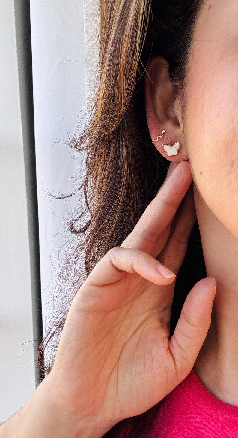 Puede incluir: Un pendiente tipo ear cuff dorado con forma de serpiente fina y sinuosa, colocado sobre un peque&ntilde;o pendiente de bot&oacute;n con forma de mariposa blanca. Las piezas de joyer&iacute;a se llevan en el l&oacute;bulo y el cart&iacute;lago, con un acabado met&aacute;lico delicado.