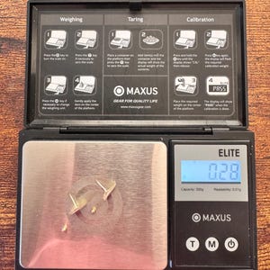 Puede incluir: Una b&aacute;scula digital negra Maxus Elite con una plataforma de pesaje de acero inoxidable que muestra 0,28 gramos. La tapa tiene instrucciones impresas para pesar, tarar y calibrar. Dos peque&ntilde;os pendientes de metal dorado descansan sobre la plataforma.