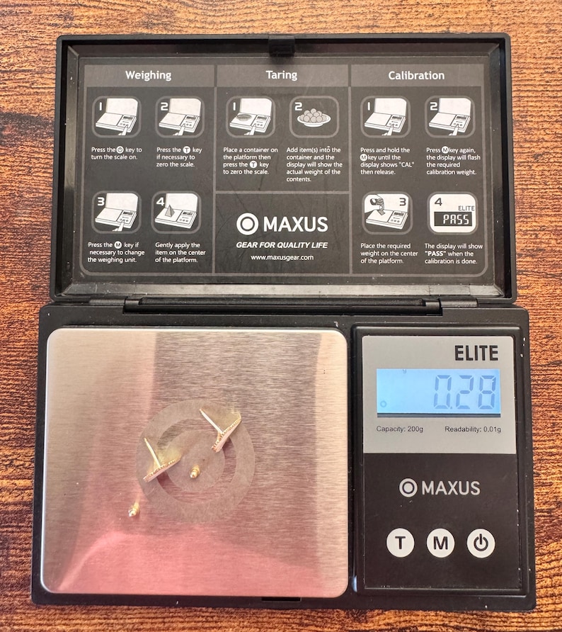 Puede incluir: Una b&aacute;scula digital negra Maxus Elite con una plataforma de pesaje de acero inoxidable que muestra 0,28 gramos. La tapa tiene instrucciones impresas para pesar, tarar y calibrar. Dos peque&ntilde;os pendientes de metal dorado descansan sobre la plataforma.
