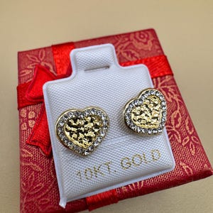 Op de afbeelding: Een paar hartvormige oorknopjes met een textuurrijk goudkleurig midden, omringd door een rand van heldere kristallen. De oorbellen zijn bevestigd op een witte displaykaart met de tekst 10KT. GOLD in gouden letters gedrukt.