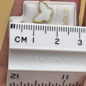 Puede incluir: Un pendiente de bot&oacute;n con forma de mariposa blanca y borde dorado con cuentas, presentado sobre una tarjeta blanca junto a una regla que muestra una anchura de unos 1,3 cm. La tarjeta incluye el texto "GOLD" y "515 MADE IN U.S.A."