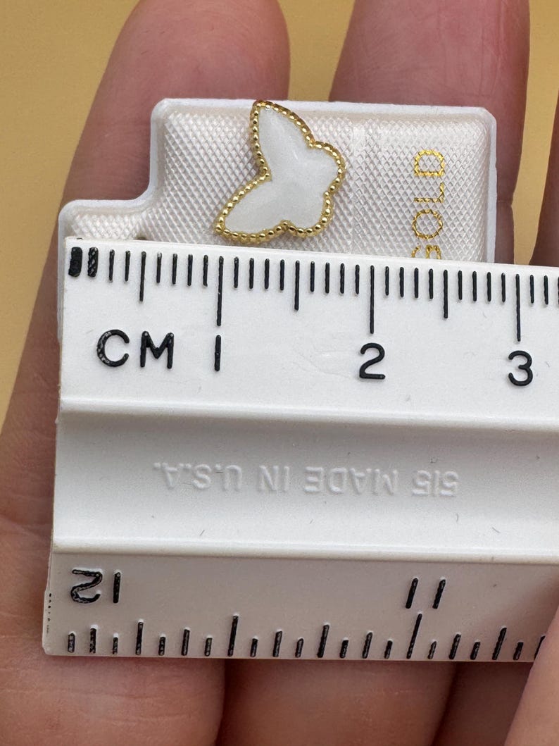 Puede incluir: Un pendiente de bot&oacute;n con forma de mariposa blanca y borde dorado con cuentas, presentado sobre una tarjeta blanca junto a una regla que muestra una anchura de unos 1,3 cm. La tarjeta incluye el texto "GOLD" y "515 MADE IN U.S.A."