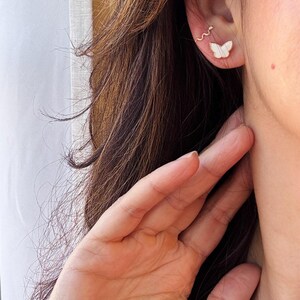 Puede incluir: Un pendiente tipo ear cuff con forma de serpiente fina y sinuosa y un pendiente de bot&oacute;n con un dise&ntilde;o de mariposa blanca con bordes dorados delicados, ambos colocados en el l&oacute;bulo y la zona del cart&iacute;lago de la oreja.
