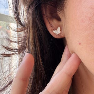 Puede incluir: Un peque&ntilde;o pendiente de bot&oacute;n con forma de mariposa que presenta una incrustaci&oacute;n blanca iridiscente con una sutil textura a rayas, enmarcado por un fino borde de metal dorado, sujeto al l&oacute;bulo de la oreja con un perno y cierre.