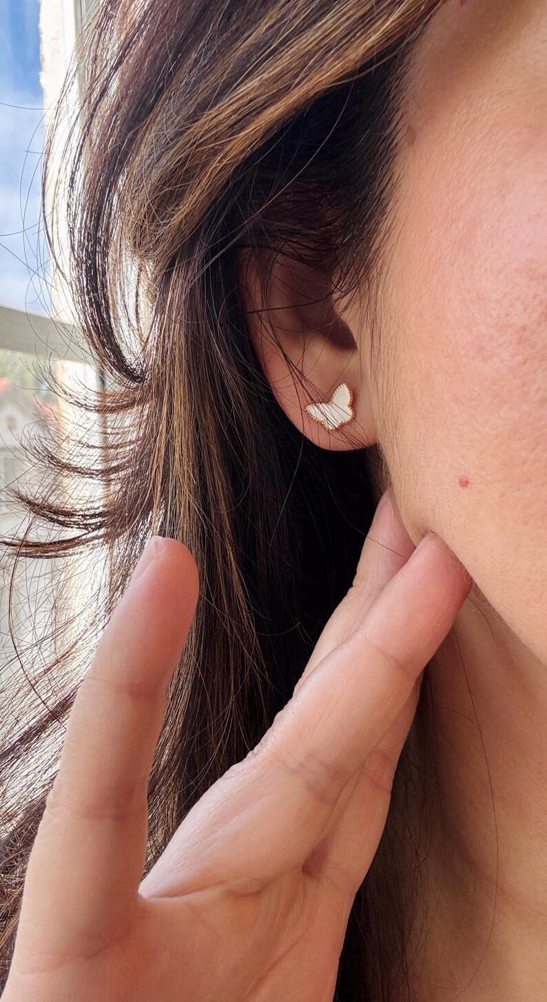 Puede incluir: Un peque&ntilde;o pendiente de bot&oacute;n con forma de mariposa que presenta una incrustaci&oacute;n blanca iridiscente con una sutil textura a rayas, enmarcado por un fino borde de metal dorado, sujeto al l&oacute;bulo de la oreja con un perno y cierre.