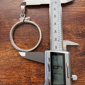 Puede incluir: Un colgante de plata con un dise&ntilde;o circular y una superficie texturizada. El colgante se est&aacute; midiendo con un calibre digital, que muestra 61,22 mm.