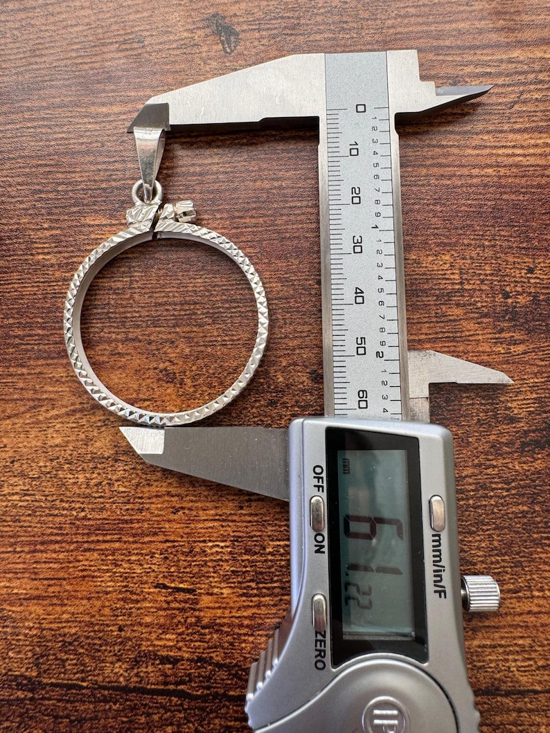 Puede incluir: Un colgante de plata con un dise&ntilde;o circular y una superficie texturizada. El colgante se est&aacute; midiendo con un calibre digital, que muestra 61,22 mm.