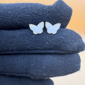 Puede incluir: Un par de pendientes de bot&oacute;n con forma de mariposa, con centros blancos y bordes dorados con cuentas. Las alas de las mariposas tienen una incrustaci&oacute;n lisa y clara rodeada por un delicado borde met&aacute;lico con un acabado texturizado.