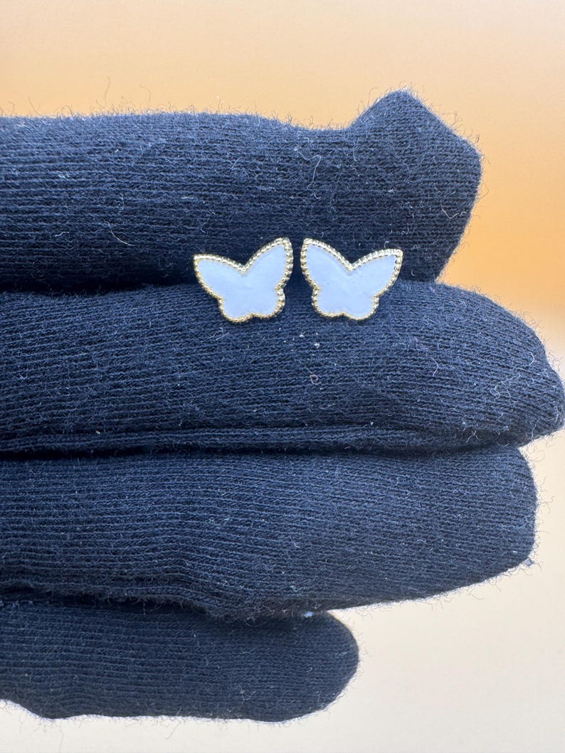 Puede incluir: Un par de pendientes de bot&oacute;n con forma de mariposa, con centros blancos y bordes dorados con cuentas. Las alas de las mariposas tienen una incrustaci&oacute;n lisa y clara rodeada por un delicado borde met&aacute;lico con un acabado texturizado.