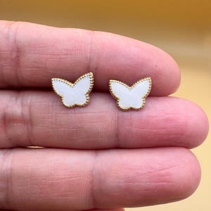 Puede incluir: Un par de pendientes de bot&oacute;n con forma de mariposa. Cada pendiente presenta una incrustaci&oacute;n central blanca rodeada por un borde fino de metal dorado texturizado. El dise&ntilde;o de la mariposa es sim&eacute;trico con alas definidas y un detalle central.