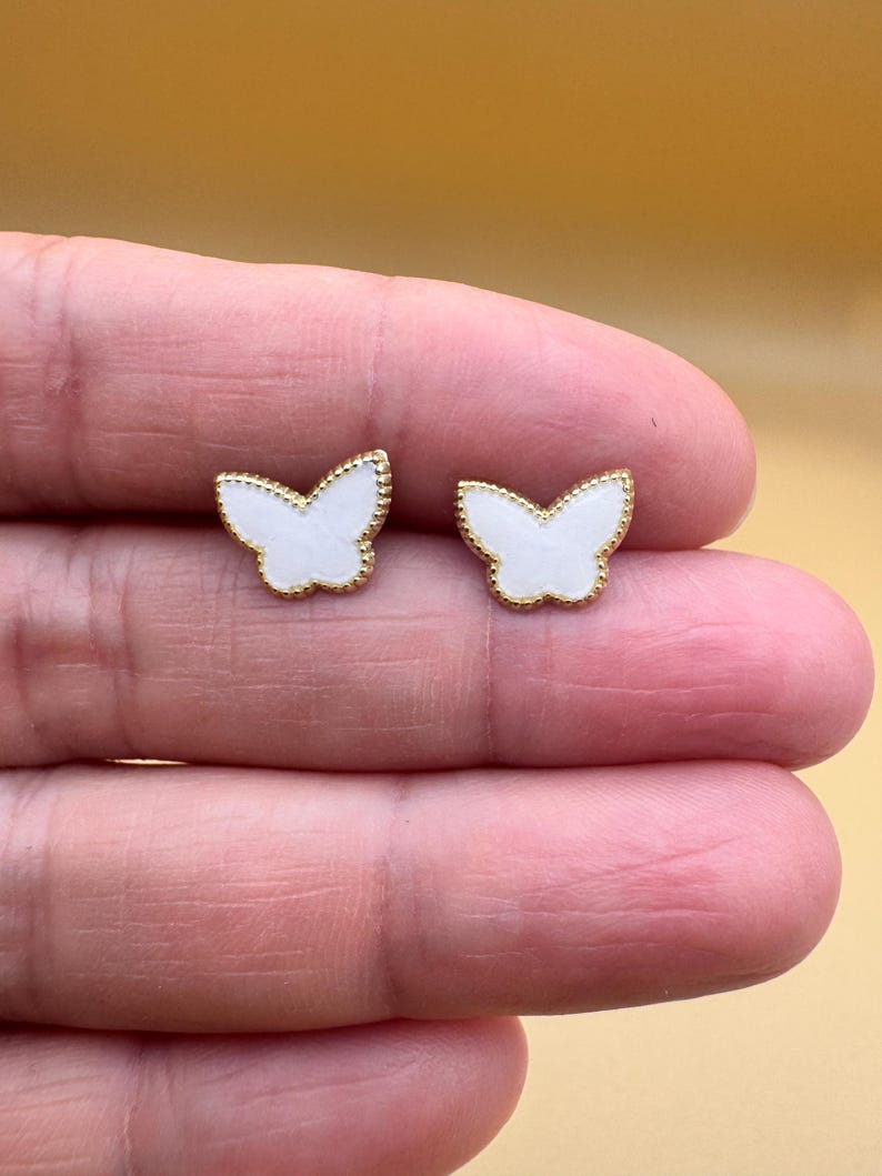 Puede incluir: Un par de pendientes de bot&oacute;n con forma de mariposa. Cada pendiente presenta una incrustaci&oacute;n central blanca rodeada por un borde fino de metal dorado texturizado. El dise&ntilde;o de la mariposa es sim&eacute;trico con alas definidas y un detalle central.