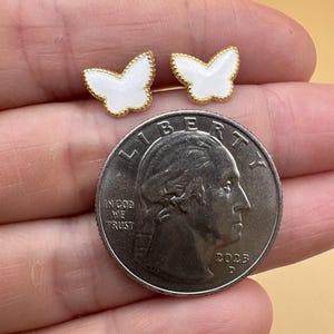 Puede incluir: Un par de pendientes peque&ntilde;os en forma de mariposa con centros de esmalte blanco y bordes festoneados en tono dorado. Los pendientes est&aacute;n colocados junto a una moneda de cuarto de d&oacute;lar estadounidense para escala, con el texto Liberty, In God We Trust y 2023.