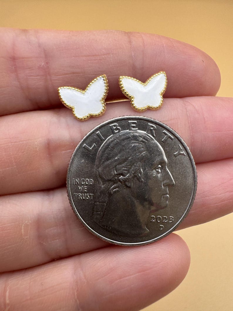 Puede incluir: Un par de pendientes peque&ntilde;os en forma de mariposa con centros de esmalte blanco y bordes festoneados en tono dorado. Los pendientes est&aacute;n colocados junto a una moneda de cuarto de d&oacute;lar estadounidense para escala, con el texto Liberty, In God We Trust y 2023.