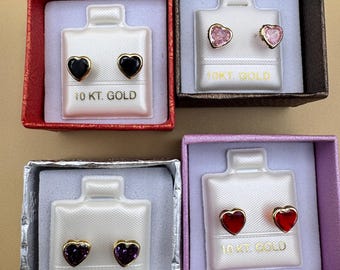 Puede incluir: Cuatro pares de pendientes de botón en forma de corazón en cajas individuales. Los pendientes tienen piedras negras, rosas, moradas y rojas engastadas en oro. Cada tarjeta de presentación blanca lleva impreso el texto 10 KT. GOLD.